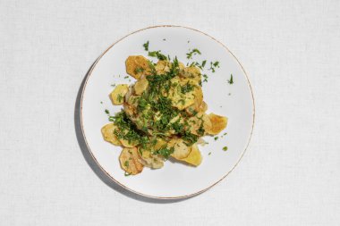 Sliced potatoes and fried in a frying pan with corn oil. Topped with finely chopped parsley. The food lies on a light, ceramic plate on a light tablecloth.