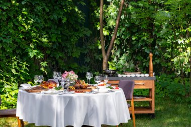On a round table with a white tablecloth are dishes from the oven and barbecue. Duck with apples, an idea with oranges, lamb ribs, steaks on light ceramic plates. The table stands on a lawn among greenery.