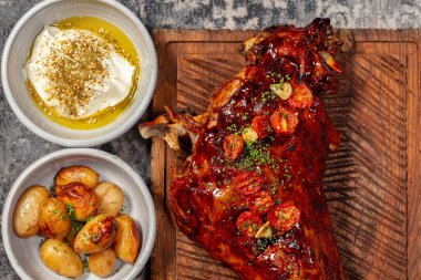 A whole leg of lamb roasted on a fire and in teriyaki sauce with fried tomatoes lies on a wooden board. Nearby are bowls of potatoes and a creamy sauce with olive oil.