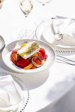 Coarsely chopped vegetables with a large piece of feta cheese, herbs and olive oil. The salad lies in a light ceramic plate on a white tablecloth. Nearby is a glass of sparkling wine, plates and cutlery.