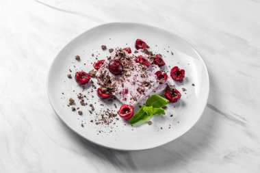 Two slices of creamy cherry ice cream lie on a light ceramic plate. Topped with chocolate chips and cherries with mint leaves. The plate sits on a marble surface.