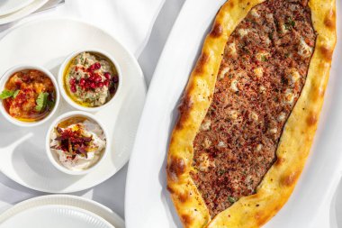 Large khachapuri with minced meat and various additives in bowls on a light ceramic plate on a white tablecloth. Additives with sauces stand side by side in light bowls.