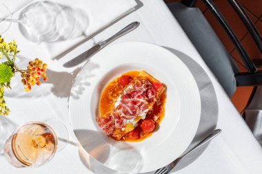 Prosciutto ile pappardelle makarna ve şarap ve krema sosuyla haşlanmış domates. Masada hafif bir seramik tabakta hafif bir masa örtüsü ile yemek. Yakınlarda bir bardak gül şarabı, bir vazo ve çatal bıçak var..