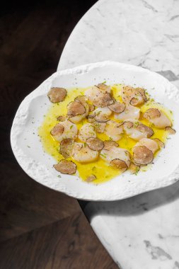 Mantarlı mantar dilimleri ve zeytinyağı ve bitkilerle karides carpaccio. Yakınlarda bir bardak köpüklü şarap ve bir şişe şarap var. Mermer tezgahta seramik tabakta yemek.. 