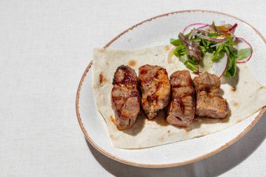 Pork kebab. Four pieces of pork kebab on a sheet of thin Armenian lavash with a mix of greens and radish salad. The food lies on a ceramic plate, on a light tablecloth.