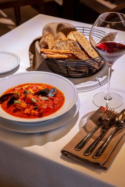 Hafif seramik tabakta deniz mahsullü domates çorbası. Çorba, yanında hafif masa örtüsü olan bir masanın üzerinde kırmızı şarap ve çatal bıçak takımı var..