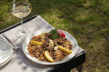 Pilaf with lamb, apples and a mix of fresh vegetable salad on a light ceramic plate. The plate is on a wooden table, next to a glass of white wine. The table is on the lawn.