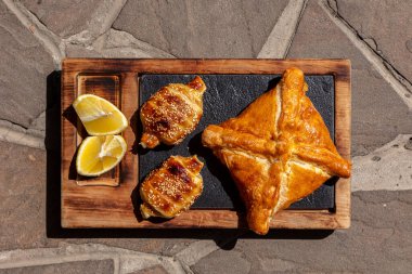 Square puff pastry khachapuri and pies with suluguni cheese and sesame seeds on top. Baking lies on a wooden board with a slate part. The plank lies on a background of stone tiles.