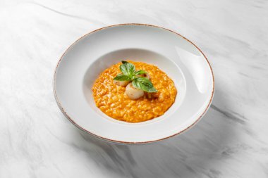 Italian dish risotto with cheddar cheese and grilled scallop, with basil leaves on top. Food in a light ceramic plate with wide margins on a marble background.