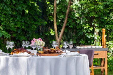 On a round table with a white tablecloth are dishes from the oven and barbecue. Duck with apples, an idea with oranges, lamb ribs, steaks on light ceramic plates. The table stands on a lawn among greenery.