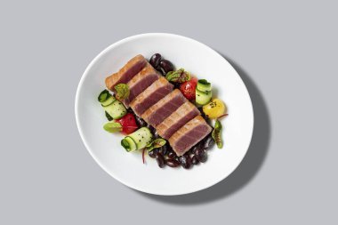 Izgara ton balığı bifteği ve porsiyon şeklinde kesilmiş. Biftek, kabak ve vişneli domatesli şarap sosunda siyah fasulyenin üzerinde yatıyor. Gri zemin üzerinde açık seramik bir tabakta yiyecek yatıyor..