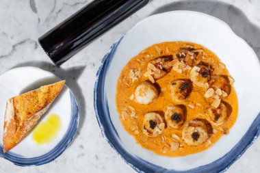 Peynir soslu siyah havyarlı ızgara deniz tarağı ve badem cipsi. Yiyecekler mermer bir tezgahın üzerindeki hafif bir seramik tabakta yatıyor. Yakınlarda bir şişe yağ ve bir tabak yağlı baget bulunur..