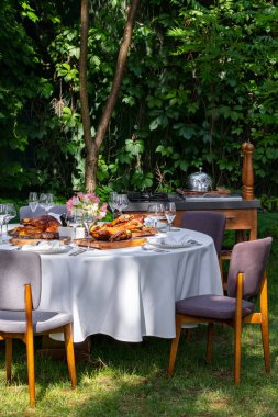On a round table with a white tablecloth are dishes from the oven and barbecue. Duck with apples, an idea with oranges, lamb ribs, steaks on light ceramic plates. The table stands on a lawn among greenery.