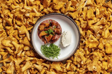 Finely chopped beef tartare topped with fried chanterelle mushrooms and chopped herbs and mushroom cream mousse. The food lies in a round ceramic plate with high sides on a pallet of fresh mushrooms.