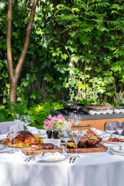 On a round table with a white tablecloth are dishes from the oven and barbecue. Duck with apples, an idea with oranges, lamb ribs, steaks on light ceramic plates. The table stands on a lawn among greenery.