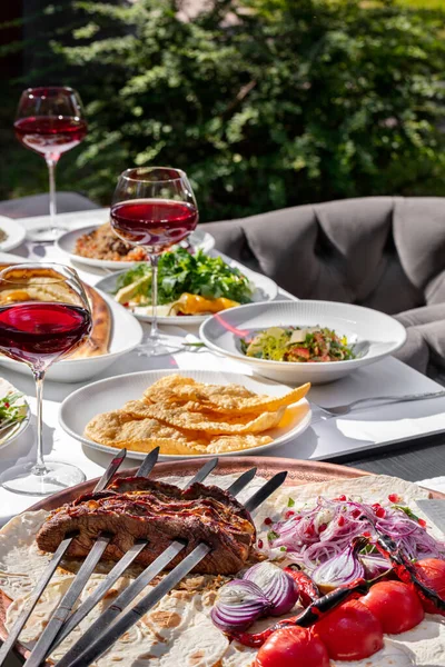 On a table with a white tablecloth is a set of dishes of Caucasian cuisine, glasses of wine, cutlery and various sauces. There are chairs around the table on the lawn. Behind is a small table with barbecue skewers.