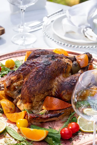 A whole turkey baked in the oven with oranges and wine. The turkey lies on a large ceramic plate with slices of orange, lemon, tomatoes and herbs. A plate with a turkey stands on a light tablecloth.