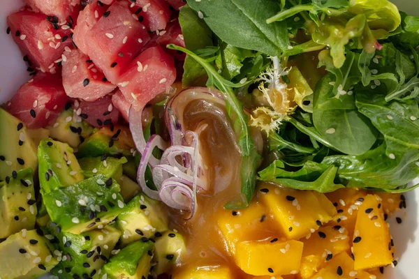 Salad with fresh tuna, avocado, herbs and mango in a light ceramic plate on a white tablecloth. Nearby is a glass of white wine and a bowl of sweet and sour sauce.