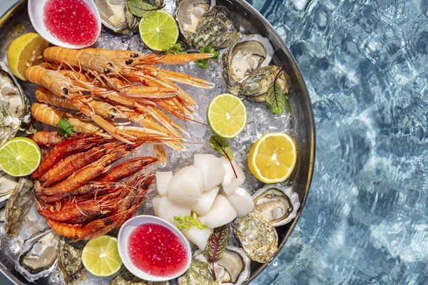 Shrimps, langoustines, scallops and oysters with various sauces and citrus fruits lie on a round metal ice tray. The tray is on the edge of the pool.