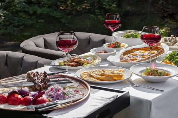 On a table with a white tablecloth is a set of dishes of Caucasian cuisine, glasses of wine, cutlery and various sauces. There are chairs around the table on the lawn. Behind is a small table with barbecue skewers.