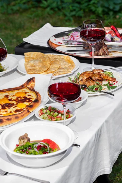 On a table with a white tablecloth is a set of dishes of Caucasian cuisine, glasses of wine, cutlery and various sauces. There are chairs around the table on the lawn. Behind is a small table with barbecue skewers.