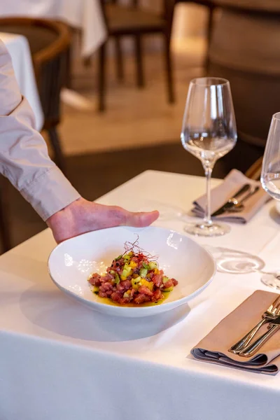 Garsonun eli masaya bir tabak ton balıklı tartar koyar. Masada boş bardak ve çatal bıçak takımı var..