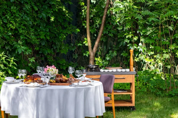 On a round table with a white tablecloth are dishes from the oven and barbecue. Duck with apples, an idea with oranges, lamb ribs, steaks on light ceramic plates. The table stands on a lawn among greenery.