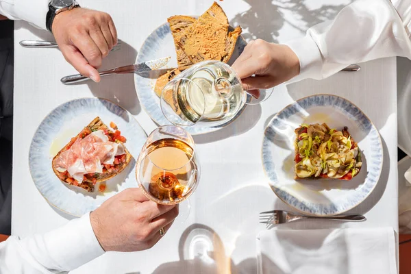 At a table with a light tablecloth with bruschettas with prosciutto and artichokes, foie gras pate and glasses of rose and white wine, a man and a woman are sitting.