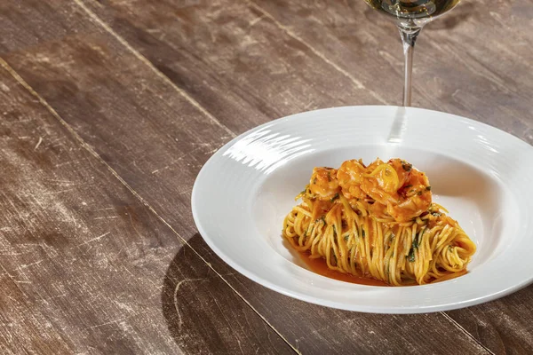トマトソースにバジルとエビのスパゲッティ 食べ物は薄いセラミックプレートにあり 隣には白ワインのグラスがあります 木製のテーブルの上に料理が置かれる — ストック写真