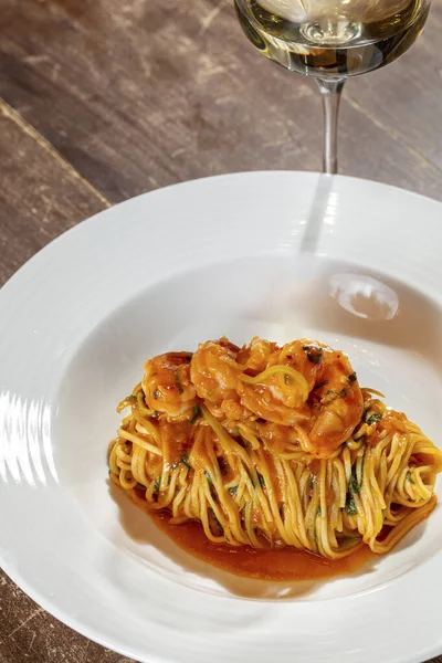 トマトソースにバジルとエビのスパゲッティ 食べ物は薄いセラミックプレートにあり 隣には白ワインのグラスがあります 木製のテーブルの上に料理が置かれる — ストック写真