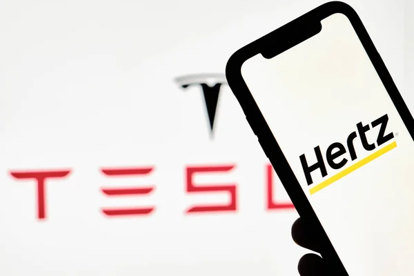 Ένα πρόσωπο κατέχει smartphone με το εμπορικό σήμα Hertz πάνω από μεγάλο λογότυπο Tesla — Φωτογραφία Αρχείου