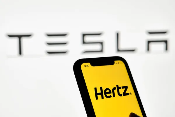 Una persona tiene un teléfono inteligente con marca registrada Hertz sobre el logotipo grande de Tesla — Foto de Stock