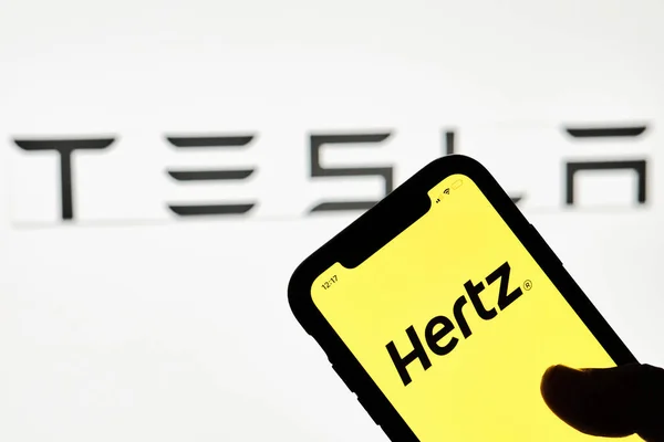 Una persona tiene un teléfono inteligente con marca registrada Hertz sobre el logotipo grande de Tesla — Foto de Stock