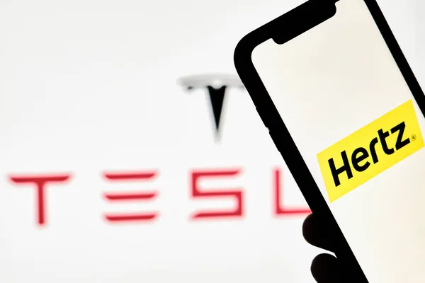 Человек держит смартфон с торговой маркой Hertz над большим логотипом Tesla — стоковое фото