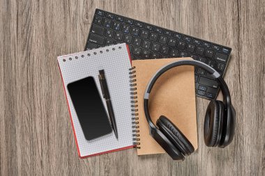 Bilgisayar, akıllı telefon, kalem, kağıt defter ve kulaklıklar ahşap masada.