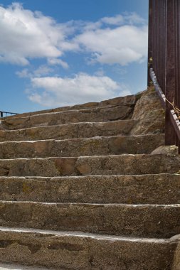 Bir tarafında demir korkuluklar olan taş merdiven yukarı çıkıyor ve çok beyaz bulutlarla mavi bir gökyüzünde bitiyor..