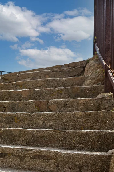 Stone staircase with iron railing on one side that goes up and ends in a blue sky with very white clouds.