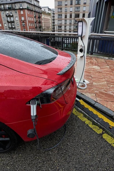 Red electric car on a rainy day connected with an adapter to an to an electric gas station. Clean energy concept as a measure to regenerate the planet from global warming and harmful CO2 emissions.