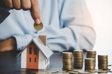 Hand putting coin in house model of coin for saving money for buying house. Savings plans for home, loan, investment, mortgage, finance and banking about house concept.