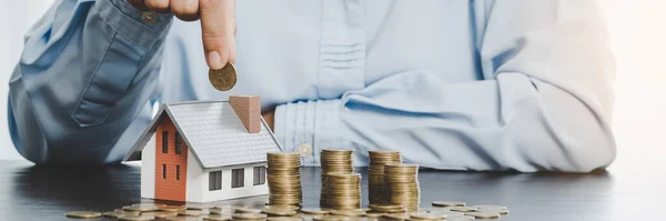 Hand putting coin in house model of coin for saving money for buying house. Savings plans for home, loan, investment, mortgage, finance and banking about house concept.