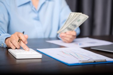 Close up young business woman using counting cash money one hundred dollar bills, checking financial documents, sitting at table with papers and tax form, managing planning budget, accounting expenses