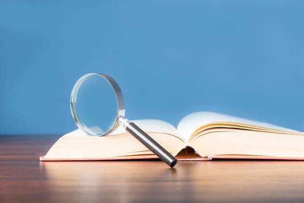 open book with magnifying glass on wooden desk in information library of school or university, concept for education,reading , study, copy space and blue background.