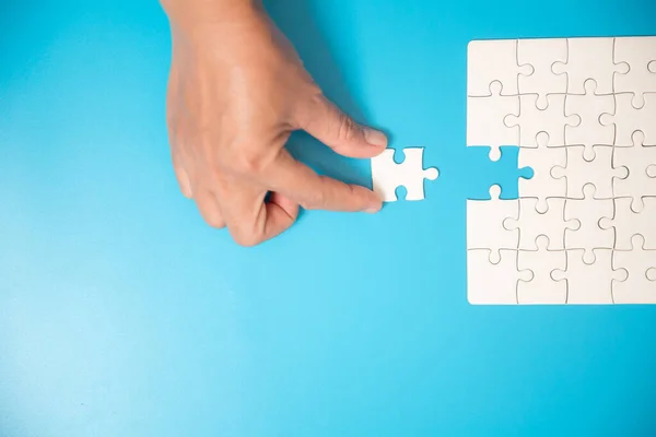 Close up hand holding and playing jigsaw game incomplete. White part of jigsaw puzzle pieces on blue background. concepts of problem solving, business, teamwork.