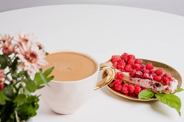 Top view of delicious piece of cake with fresh raspberries in golden plate and cup of delicious coffee next to cake on white table.  Blurry chrysanthemums in the foreground. Sweet concept.  Place for your text.