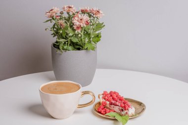 Luxurious cup of coffee and delicious piece of cake with fresh raspberries next to cup of coffee on white table. Beautiful pink chrysanthemums in grey pot in background. Light grey background. Place your text.