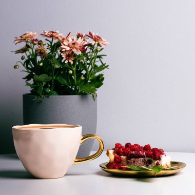Luxurious cup of coffee and delicious piece of cake with fresh raspberries next to cup of coffee on white table. Beautiful pink chrysanthemums in grey pot in background. Light grey background. Place your text.