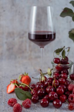 Appetizing, fresh, juicy, ripe grape, strawberry and raspberry assortment on light gray background. Blurry glass of red wine and transparent jar with cherries in background. Greeting card. Post card. Place for your text.