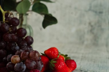 Appetizing, fresh, juicy, ripe grapes and strawberry assortment on light gray background. Glass vase with greenery in the background. Excellent image for banners and advertisements. Greeting card. Post card. Place for your text.