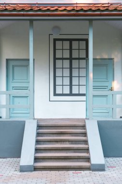 Dekoratif ahşap korkulukları ve ön planda büyük bir taş merdiveni olan yenilenmiş beyaz bir binanın ön cephesi..