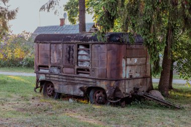 Meadow 'da büyük bir ladin altında park edilmiş eski model bir karavan. Arka planda eski çatı, yeşillik ve gökyüzü.